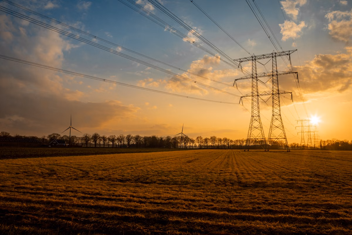 Overhead electricity transmission pylons and power lines crossing a flat field at sunset, with wind turbines visible in the distance.