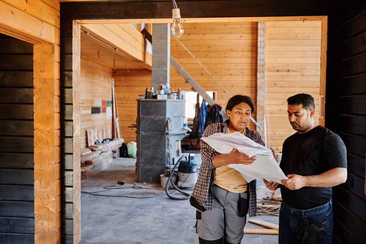 A tradeswoman holding a set of plans talks with a man inside a timber-clad house under renovation, with tools and materials in the room behind them.