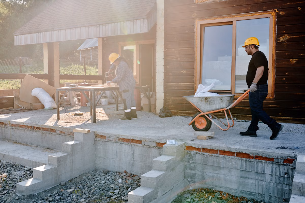 Two workers in hard hats on a small residential building site, one pushing a wheelbarrow past a timber-clad house and the other working at a trestle table.