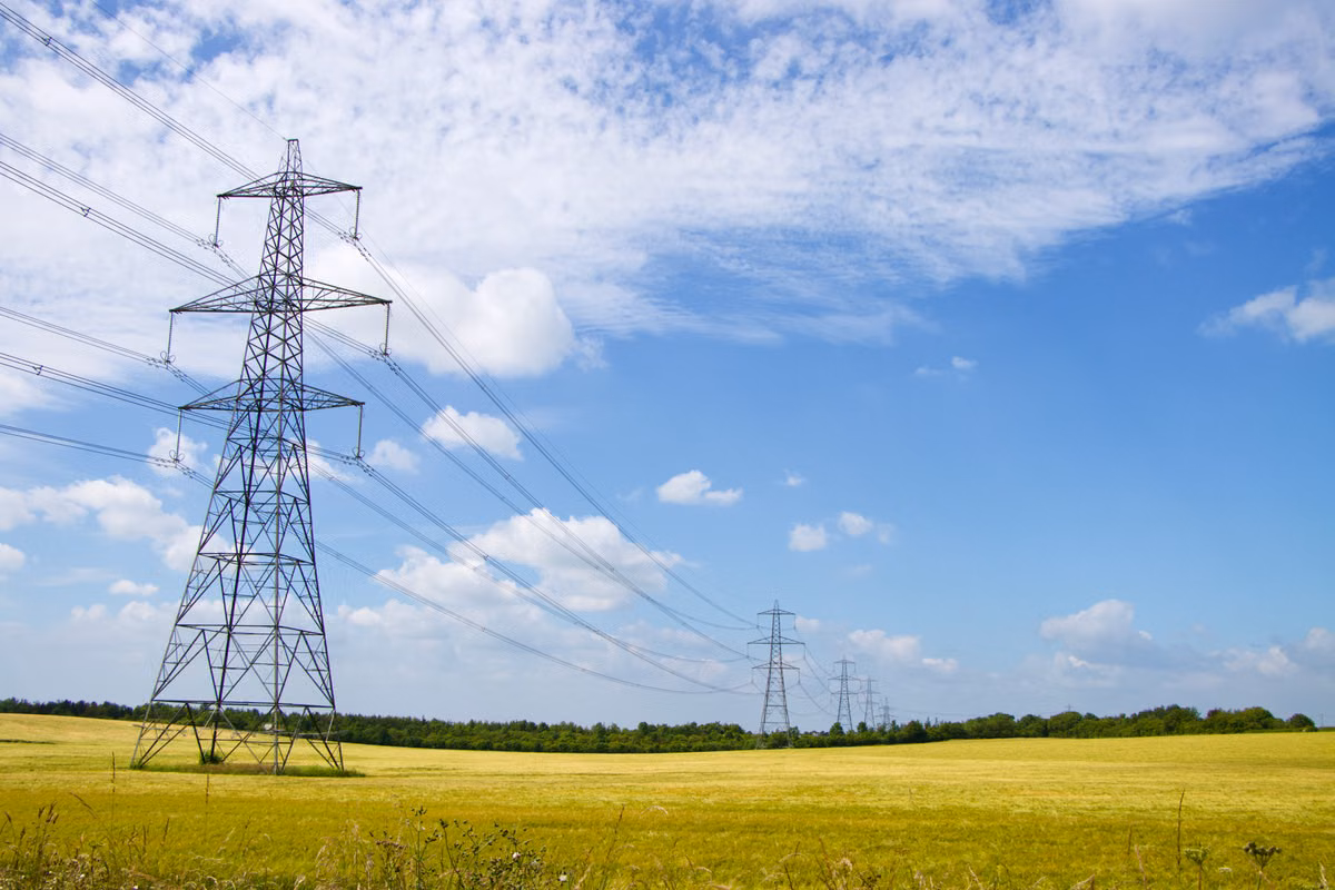 A line of electricity pylons carrying overhead power lines across open farmland under a partly cloudy blue sky.