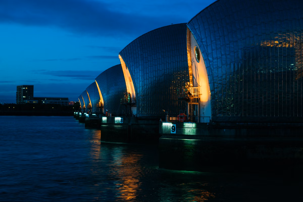 The steel-clad gate piers of a river flood barrier lit up at dusk, with numbered piers above the water.