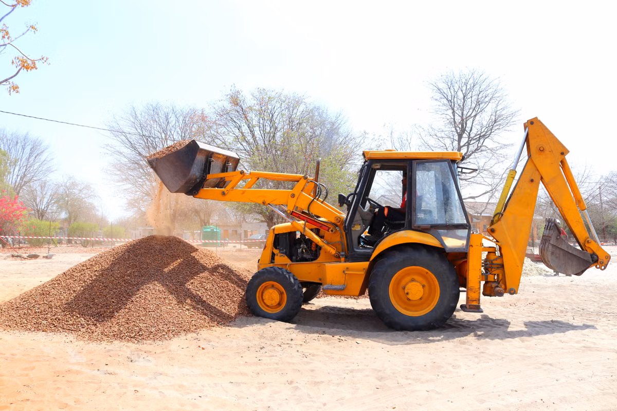 A yellow backhoe loader with no visible branding tips gravel onto a pile on a sandy site.