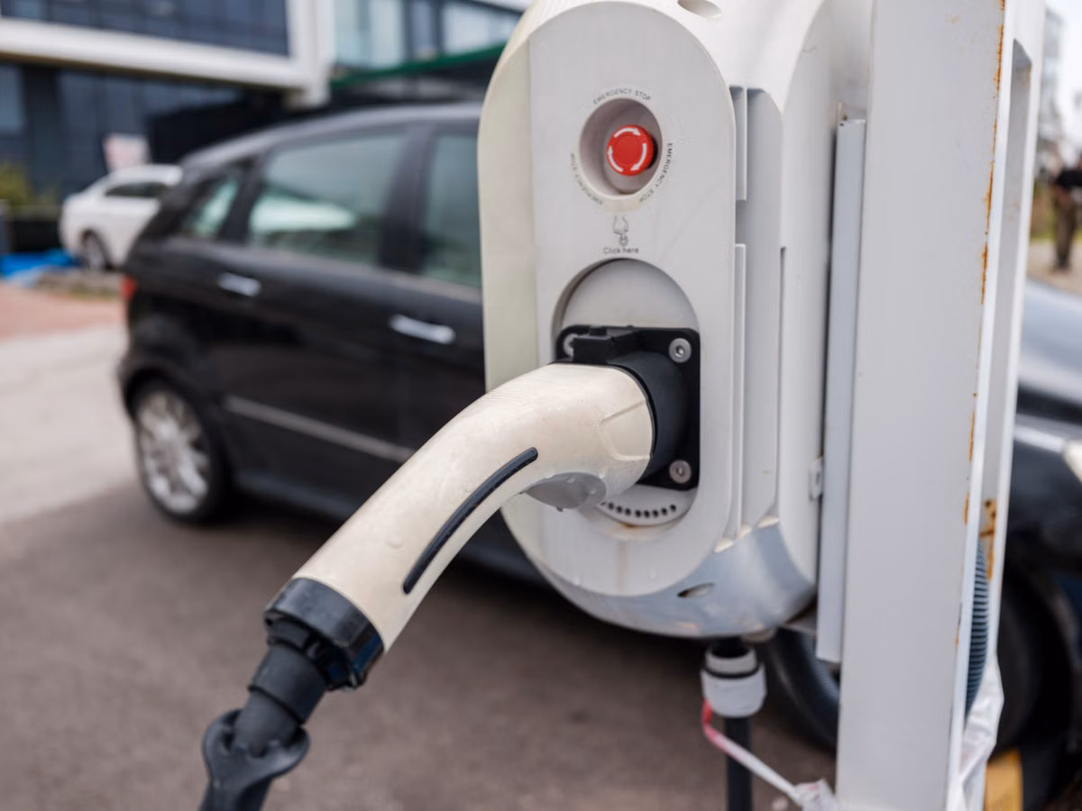 Close-up of an electric vehicle charging unit with its connector plugged in, with a parked car in the background.