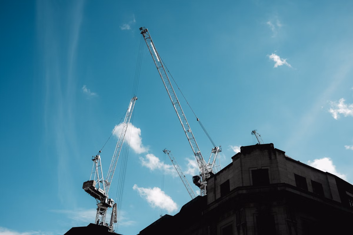Several white tower cranes rise against a blue sky behind the top of a stone-faced city building.