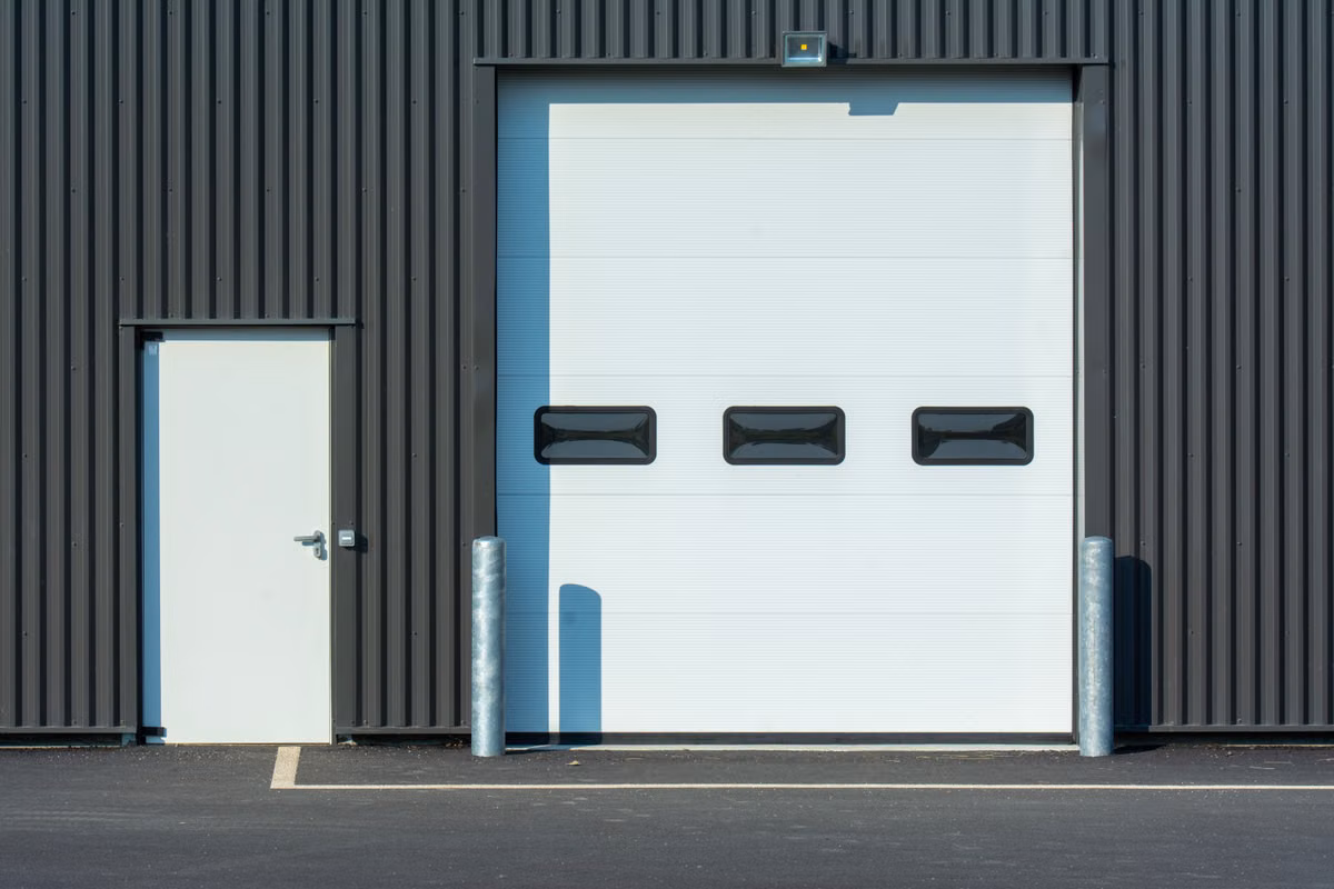A dark grey clad industrial unit with a white sectional loading door, a white personnel door and two steel bollards beside a tarmac yard.